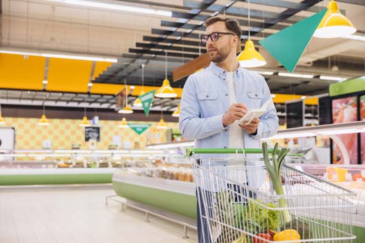 Un hombre con gafas empuja un carrito de supermercado lleno de verduras mientras mira a un lado.