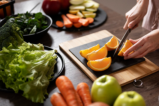 Manos cortando naranjas frescas en una tabla junto a verduras verdes para la preparación de una dieta saludable y equilibrada.