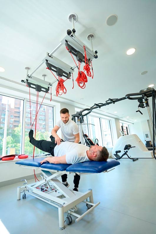 Un fisioterapeuta joven asiste a un hombre de mediana edad que yace boca arriba en una camilla de fisioterapia roja, mientras realiza un ejercicio de elevación de pierna suspendida con un sistema de cuerdas de rehabilitación en un gimnasio médico moderno con grandes ventanas.