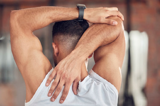Vista posterior de un hombre con camiseta blanca estirando el tríceps y el hombro derecho detrás de la cabeza en un gimnasio. Lleva un reloj inteligente deportivo en la muñeca izquierda.