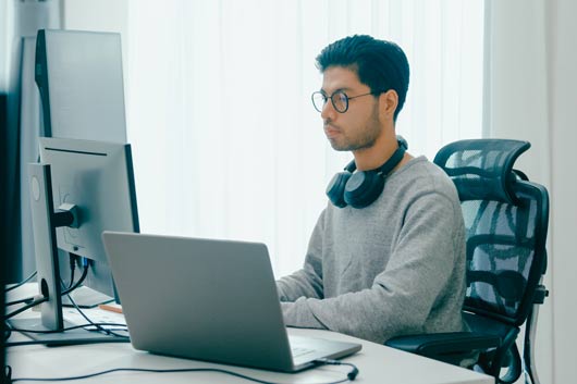 Joven programador o desarrollador web con gafas y auriculares, concentrado trabajando en un portátil y múltiples pantallas en una oficina luminosa, realizando tareas de codificación o análisis de datos.