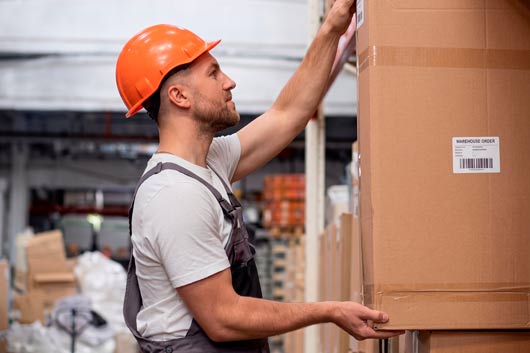 Trabajador de almacén con casco de seguridad naranja y uniforme, levantando o colocando una caja de cartón grande en una estantería, en un almacén con inventario apilado.