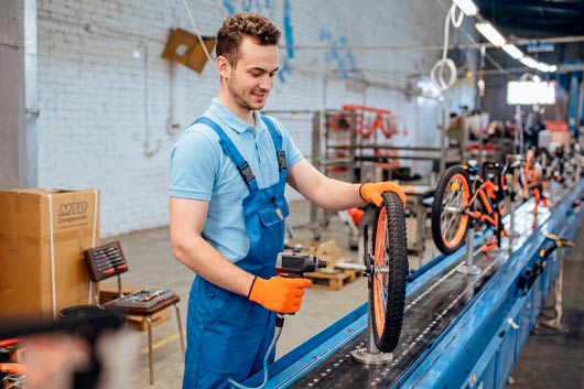 Obrero en una fábrica de bicicletas, vistiendo uniforme de trabajo y guantes, sonriendo mientras ensambla la rueda de una bicicleta naranja en una línea de producción industrial, utilizando herramientas eléctricas.