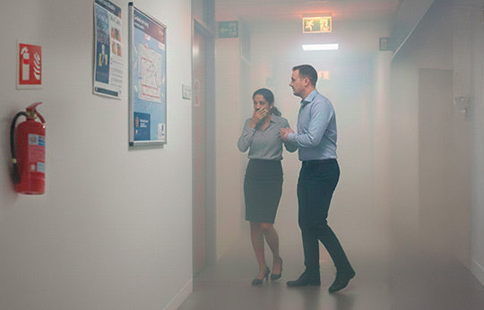 Personas evacuando una oficina llena de humo durante un incendio, con un extintor y señal de salida de emergencia.
